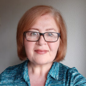 Susan Littlemore, PR Manager, a middle-aged white female with ginger hair in a bob, with big black glasses, wearing a blue patterned blouse.