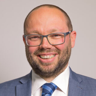 Gareth McNab - Head of External Affairs, Christians Against Poverty, smiling at the camera. He is a white male with very short hair, glasses and is wearing a grey suit with white shirt and stripy blue tie.