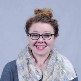 Hannah White, a white lady with tied-up auburn hair, wearing black glasses, bright lipstick and a scarf.