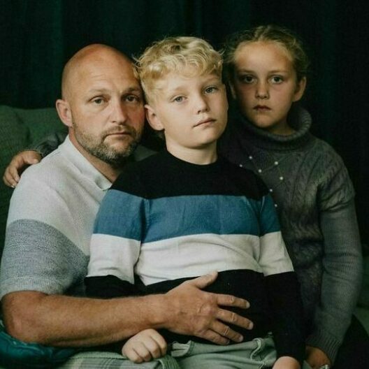 Parent sat with his two children, the boy is sat on his lap. All three are looking at the camera with sadness