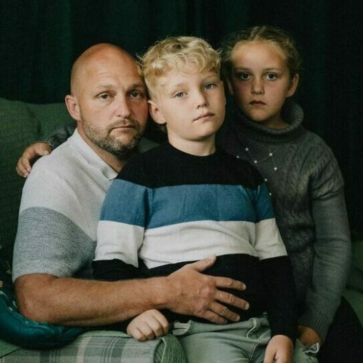 Man sat on the sofa with his two children, all three are looking into the camera