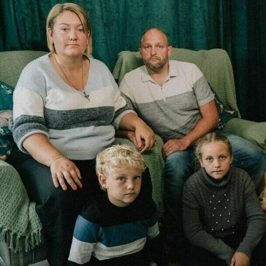 A family of two parents and two children, sat on a sofa next to a Christmas tree, looking at the camera with sadness