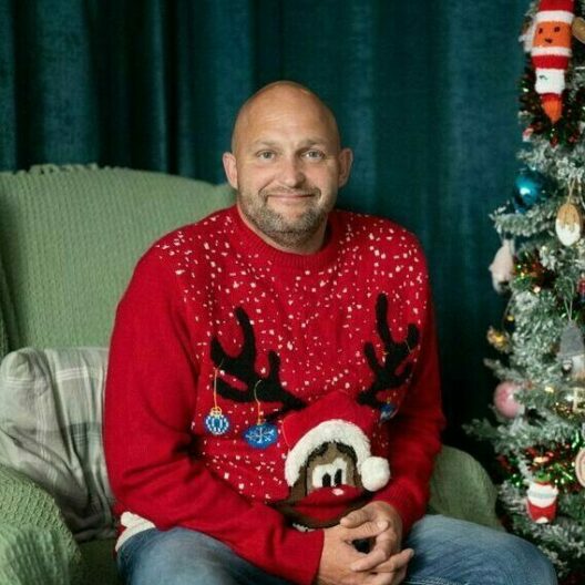 Man sat on a sofa wearing a Christmas jumper, smiling at the camera