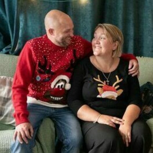 A couple sat on the sofa hugging, both are wearing Christmas jumpers and sat next to a Christmas tree