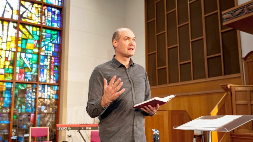 A man holding an open bible, giving a talk at the front of a church