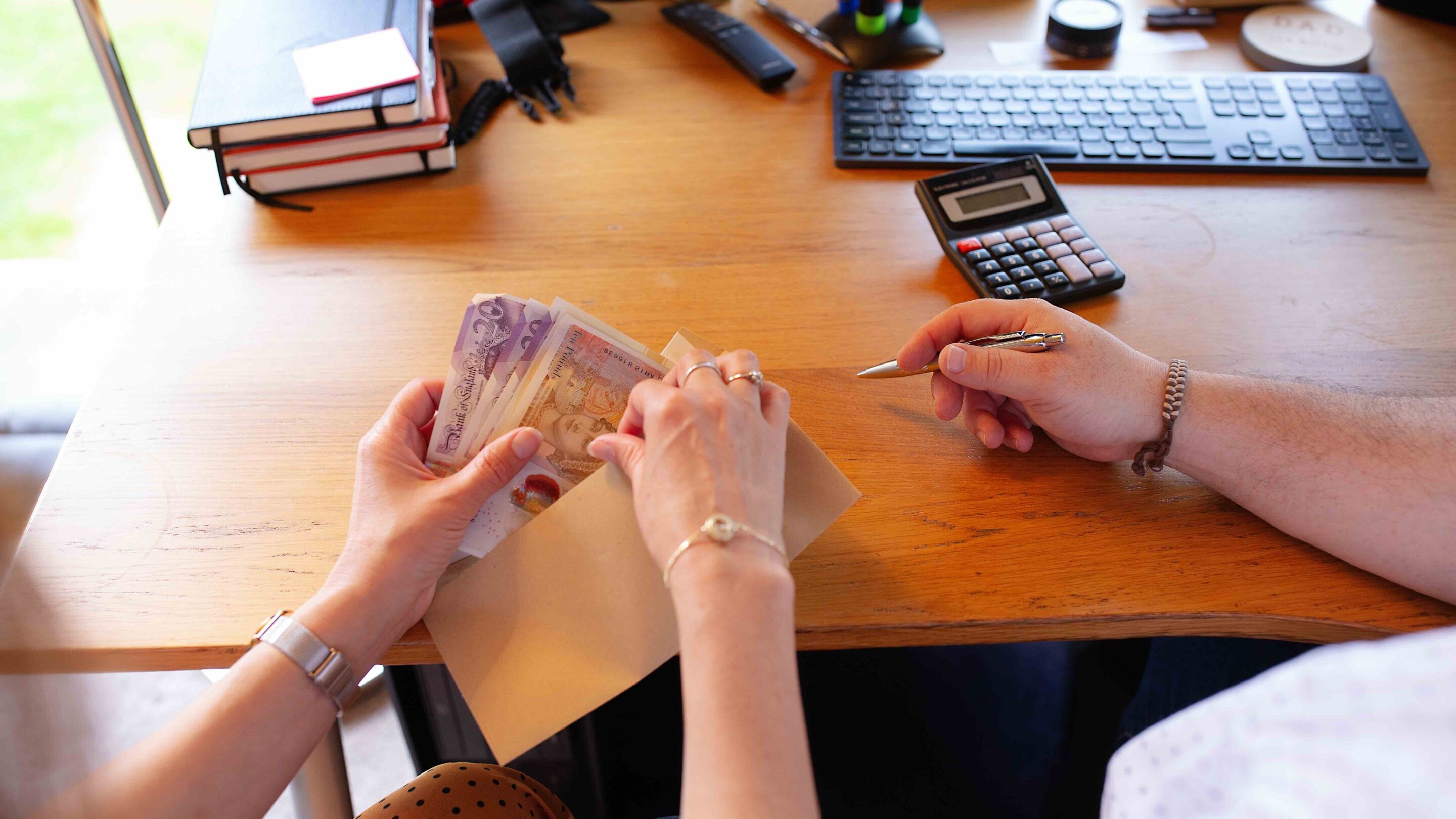 female hands holding a long brown envelope with a handful of £20 and £10 notes, male hand next to her holding a silver pen with a calculator placed next to him.
