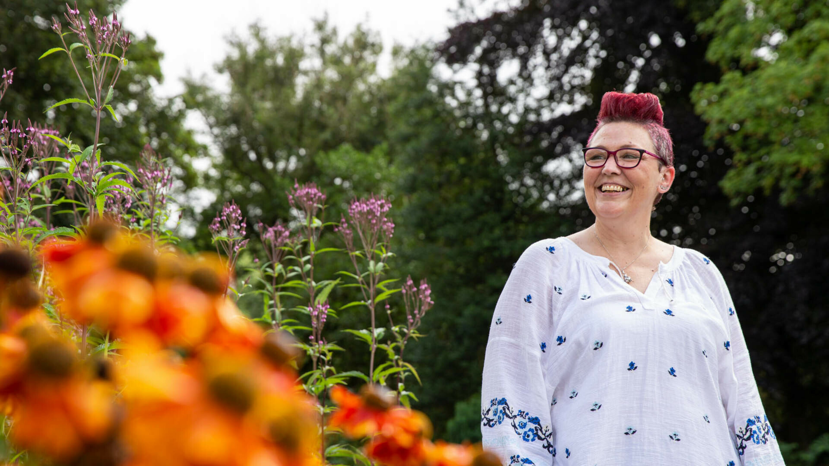 Smiling woman with dyed red hair looks towards a foreground of bright orange flowers