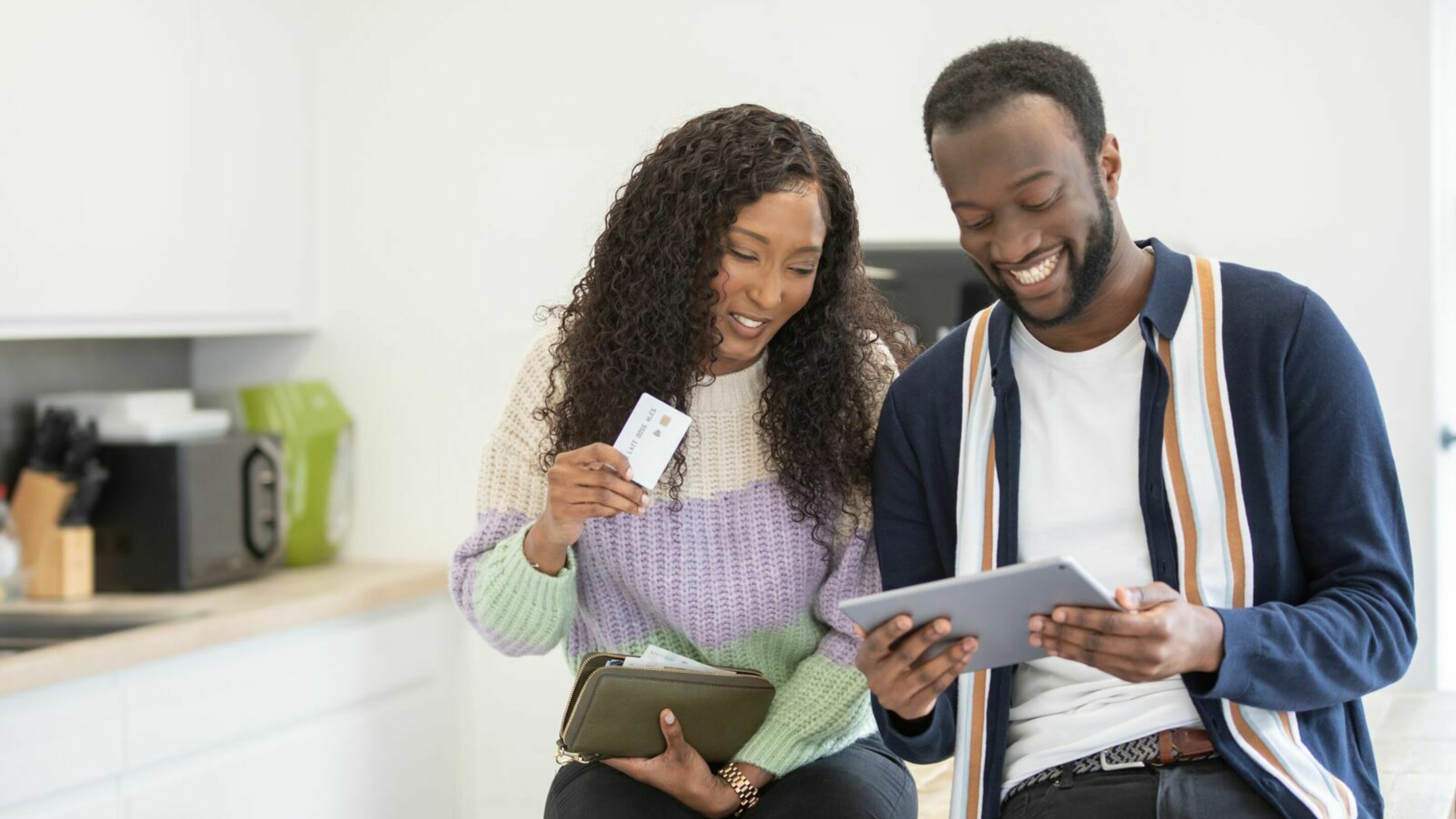 A young black couple, sitting in their kitchen. She is holding a card and purse, and he's sat next to her looking at a computer tablet. They're both smiling candidly.