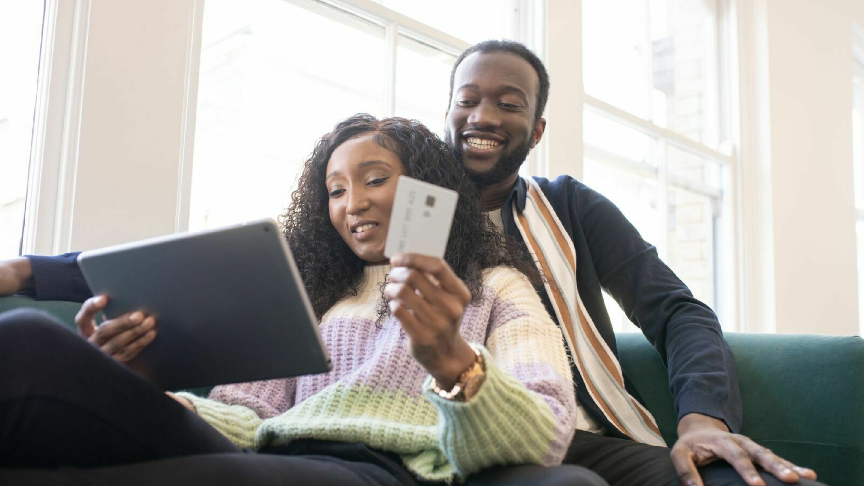 A black man and woman sat together on the sofa. She is holding a tablet and a card, and they're looking at the tablet together