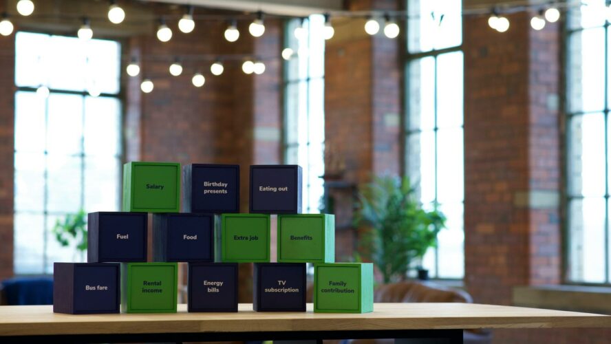 A series of green and navy cubes, stacked in a pyramid with the cubes in no particular order. Each of the navy cubes mentions a cost: food, fuel, birthday presents, eating out, bus fare, energy bills, and TV subscription. Each of the green cubes contains a means of income: benefits, extra job, salary, rental income and family contribution.