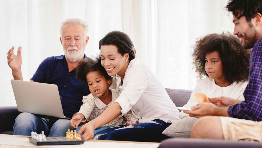 An older man sits on a sofa, with a younger woman and her young daughter. Next to them is a young girl with curly hair and her dad. They are looking at laptop and there is a game of chess on the table.