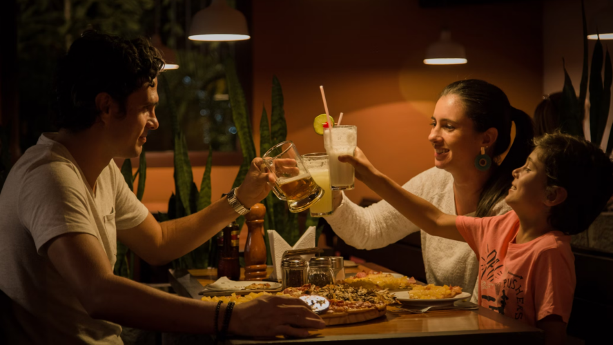 A dad, mum and boy out for dinner, holding their drinks in a 'cheers' motion over a dinner of pizza.