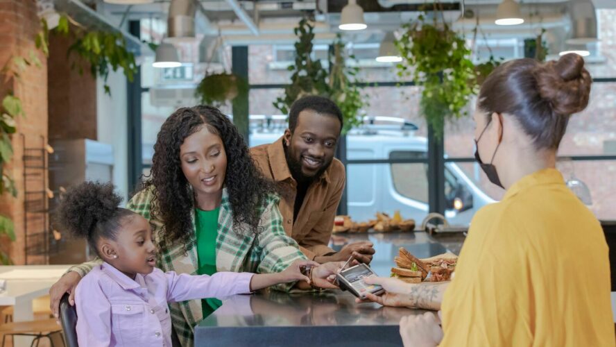 At a cafe: a young black family of three are at the counter where a white lady with a mask on is offering them the card payment machine. The young girl is holding out her hand with a card to pay.