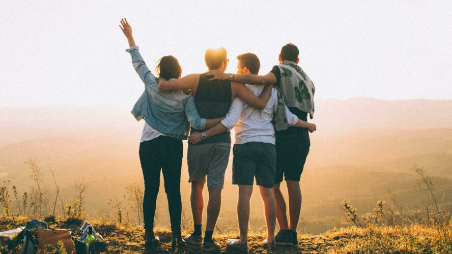 Four friends, taken from behind. They're stood on the edge of a hill, with hills and sunshine in front of them. The four have their arms around each other, enjoying the view together, as if they're enjoying the victory and adventure. Photography by Helena Lopes.