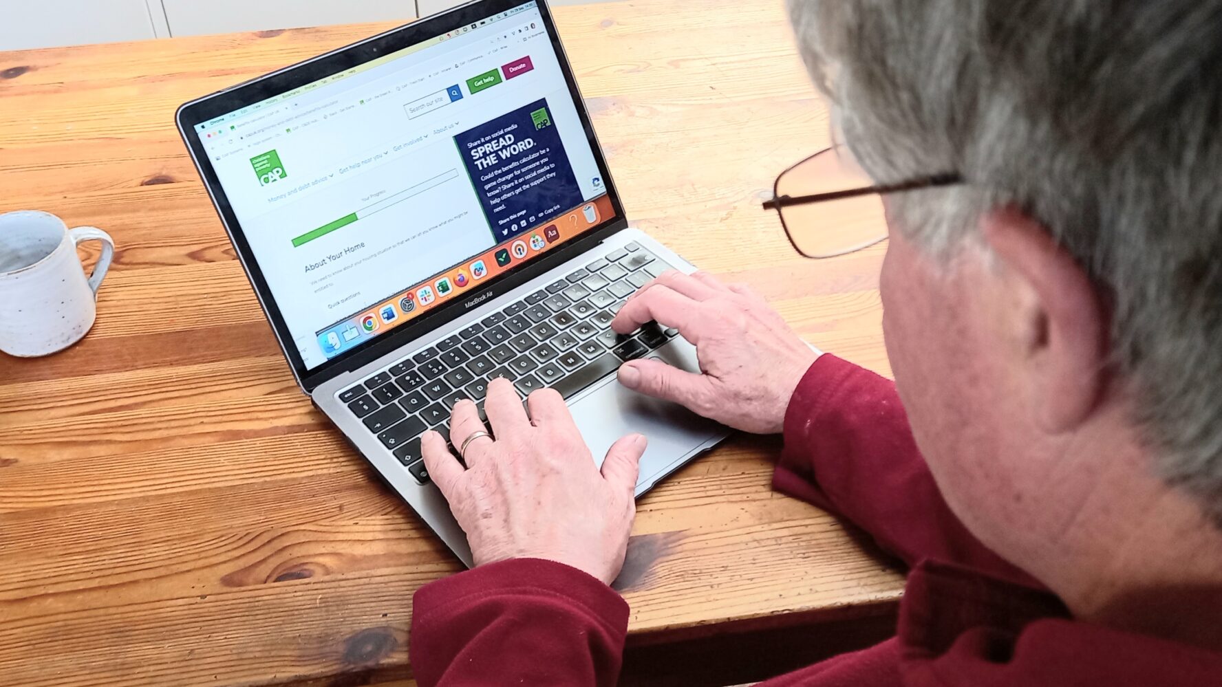 A laptop on a table, open on the benefits calculator page. A man is sat completing the calculator online.