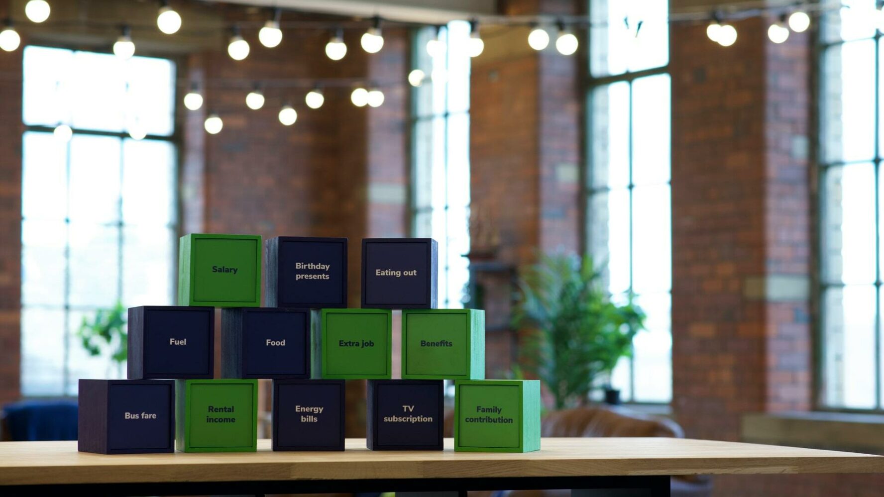 A series of green and navy cubes, stacked in a pyramid with the cubes in no particular order. Each of the navy cubes mentions a cost: food, fuel, birthday presents, eating out, bus fare, energy bills, and TV subscription. Each of the green cubes contains a means of income: benefits, extra job, salary, rental income and family contribution.