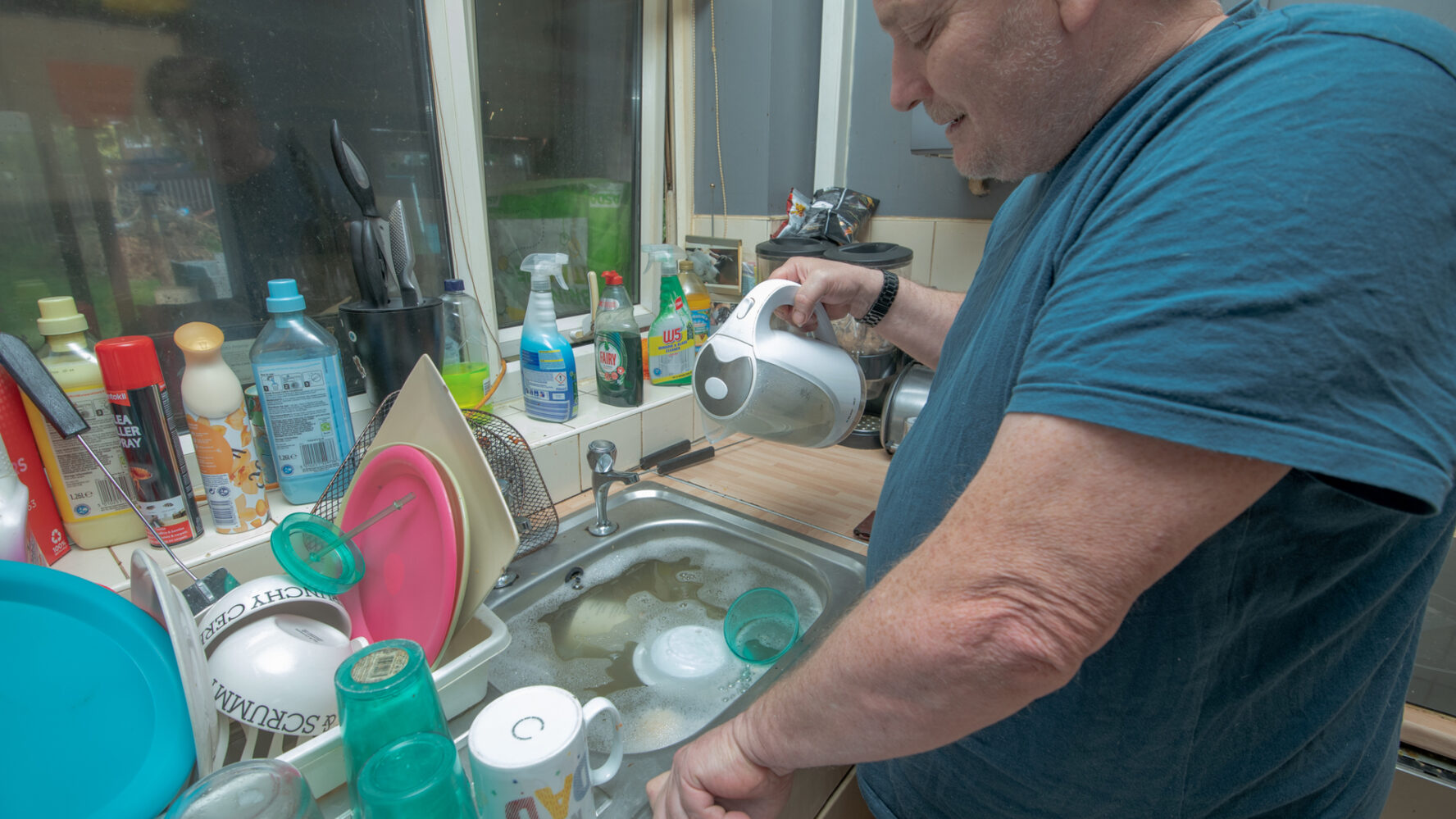 Older male pouring water from a kettle into his sink which is full of cups and bowls.