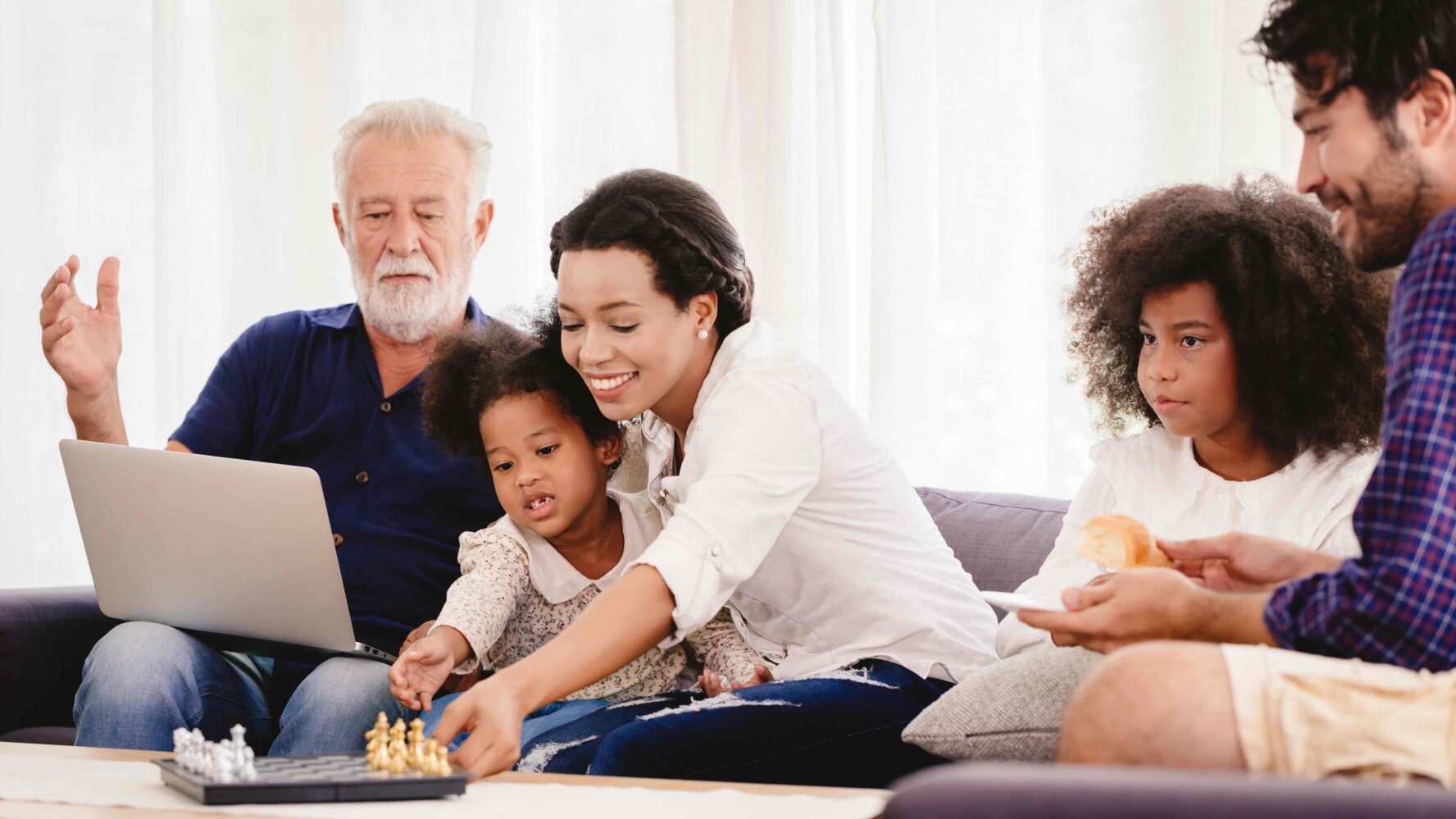 An older man sits on a sofa, with a younger woman and her young daughter. Next to them is a young girl with curly hair and her dad. They are looking at laptop and there is a game of chess on the table.
