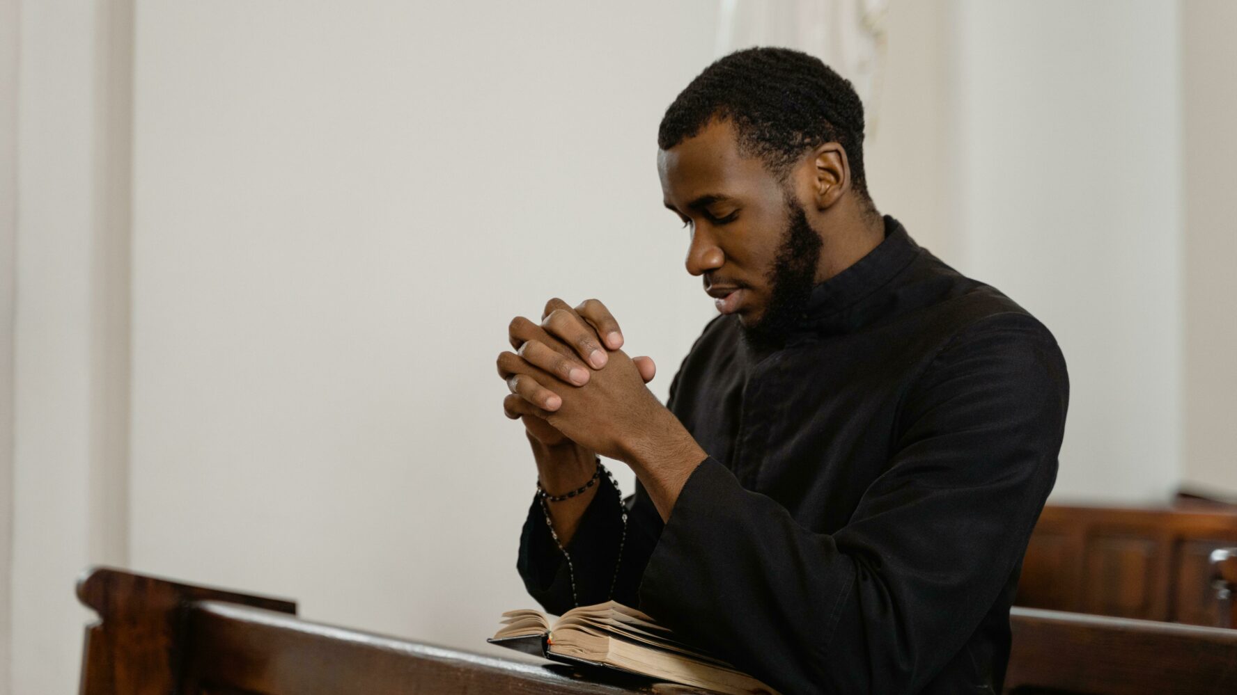 A young black man sat at a church pew, with his hands clasped together and eyes shut, praying. Photography by Mart Production.