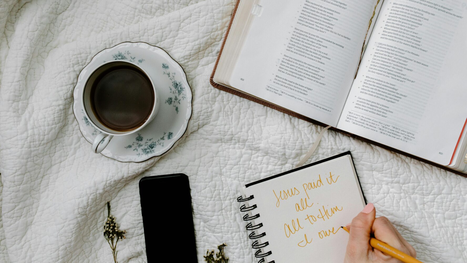 An open Bible, cup of tea, phone and notebook on a white blanket