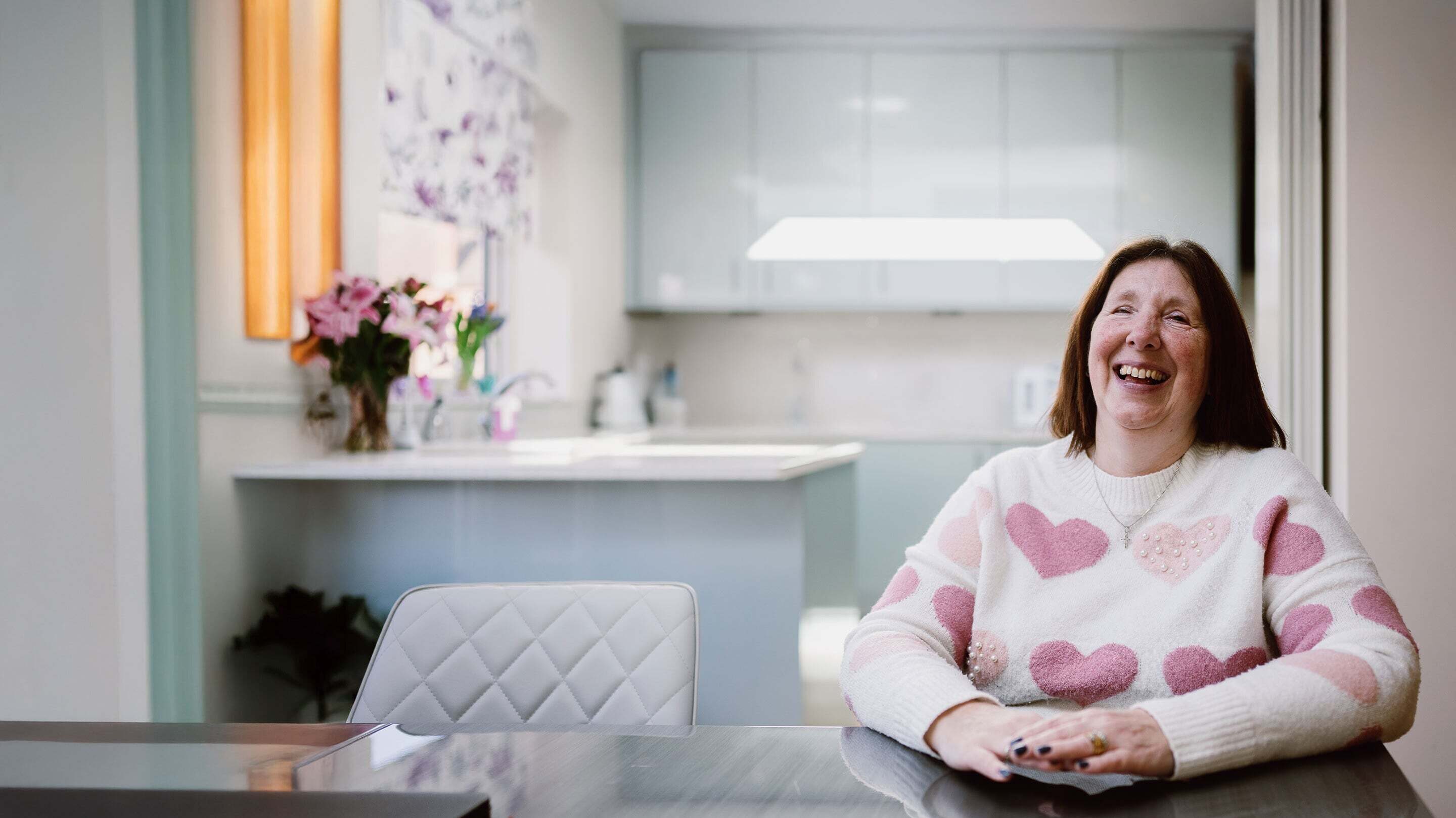 Angela, mum of three, with shoulder length straight brown hair, and a white jumper with pink hearts. She is laughing and very happy.