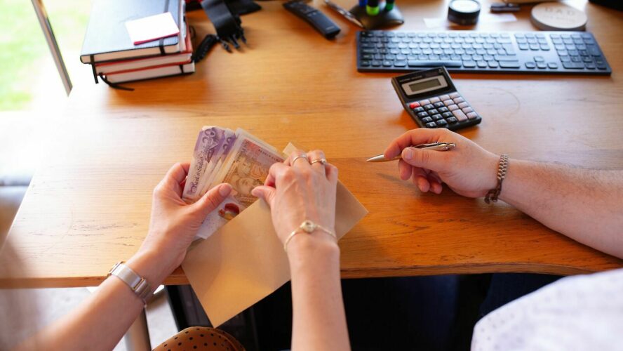 Two people are sat at a wooden table or desk. A women's hands are holding some £20 notes which she's putting into an envelope. The man is holding a pen.