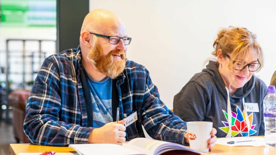 Life skills attendee, Kieron, a white man with a bald head, glasses and beard, smiling widely during one of the life skills sessions. He's sat at the table with a workbook open in front of him.