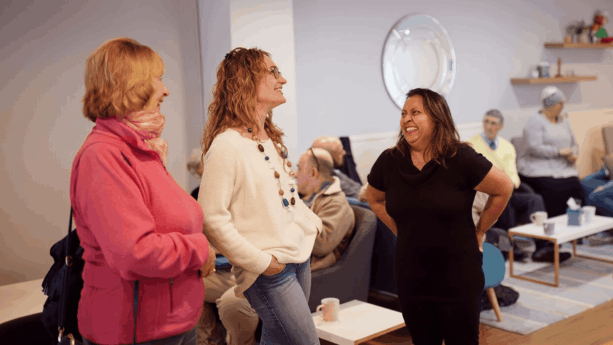 Three ladies of different ages and nationalities laughing and chatting, in a warm and inviting church community space filled with other people in the background.
