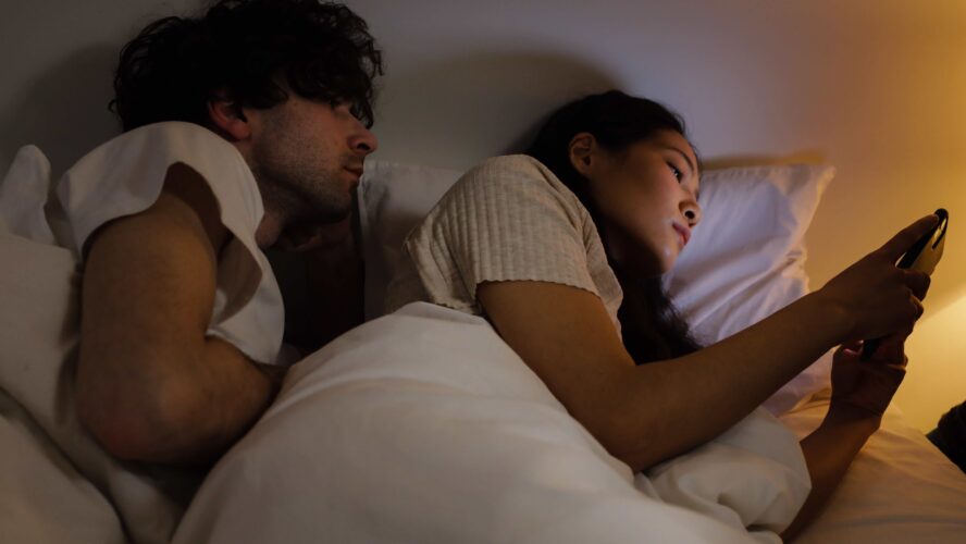 Male and female lying in bed together looking at a phone screen.