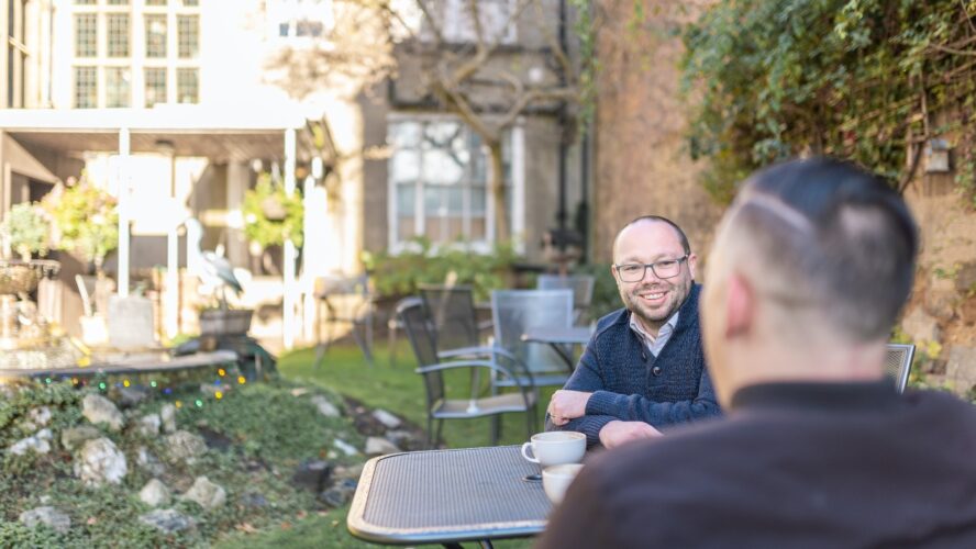 Two older men talking and smiling to each other outside in the garden with a hot drink in a cup.