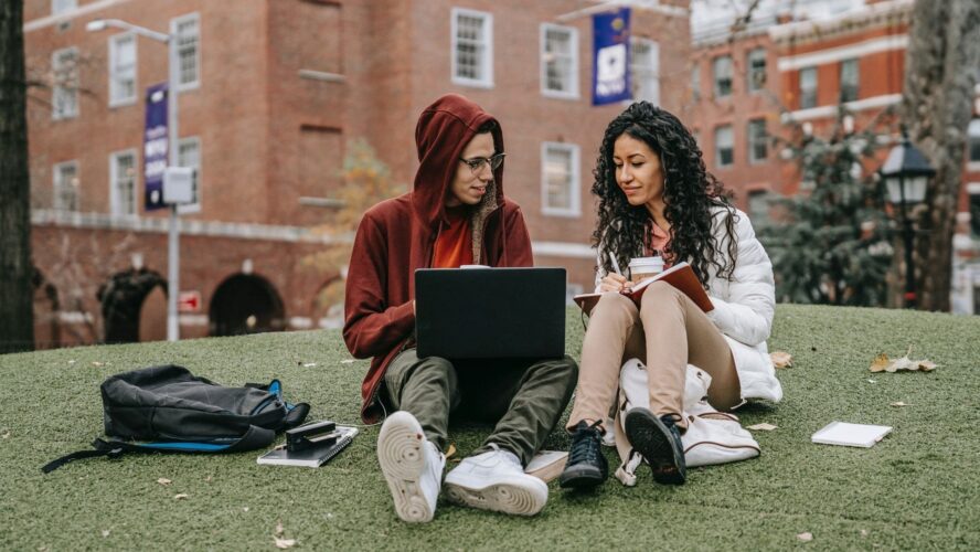 Two students sat on the grass with the orange brick university building behind them. The male, on the left, is holding a laptop, showing something to the female (on the right), who is taking notes in a notebook.
