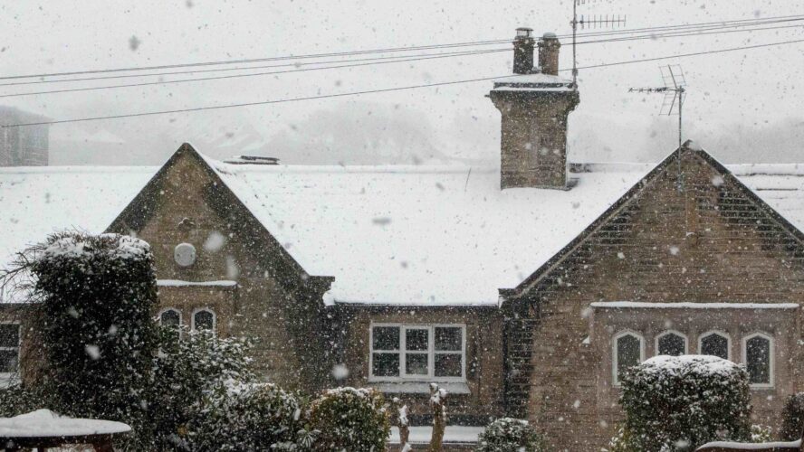 A wintery landscape of a large house with a chimney covered in snow, there are picnic tables in front.