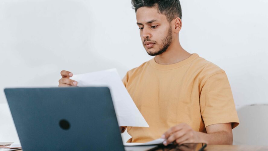 Young male, looking at piece of paper and typing on his laptop.