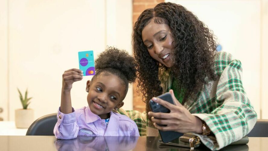 Mum and daughter smiling looking at a phone. The daughter is holding up a bank card in her right hand.