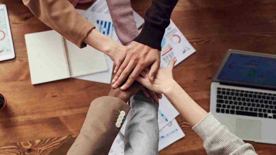 From above: six people doing a 'hand stack', putting their hands into the middle of a circle. Underneath them are documents that refer to numbers and finances.