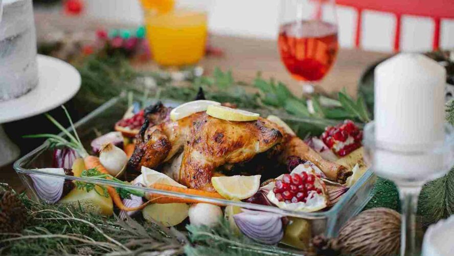 A Christmas table set up with candles, sprigs of foliage and cups filled with drinks. In the middle is a roasted chicken in a pyrex dish surrounded by onions and other vegetables.