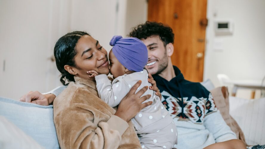 A mum and dad sat on their sofa, smiling at their baby