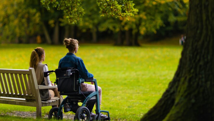 Picture of a woman sat in a wheelchair in a park, sat next to another person on a park bench