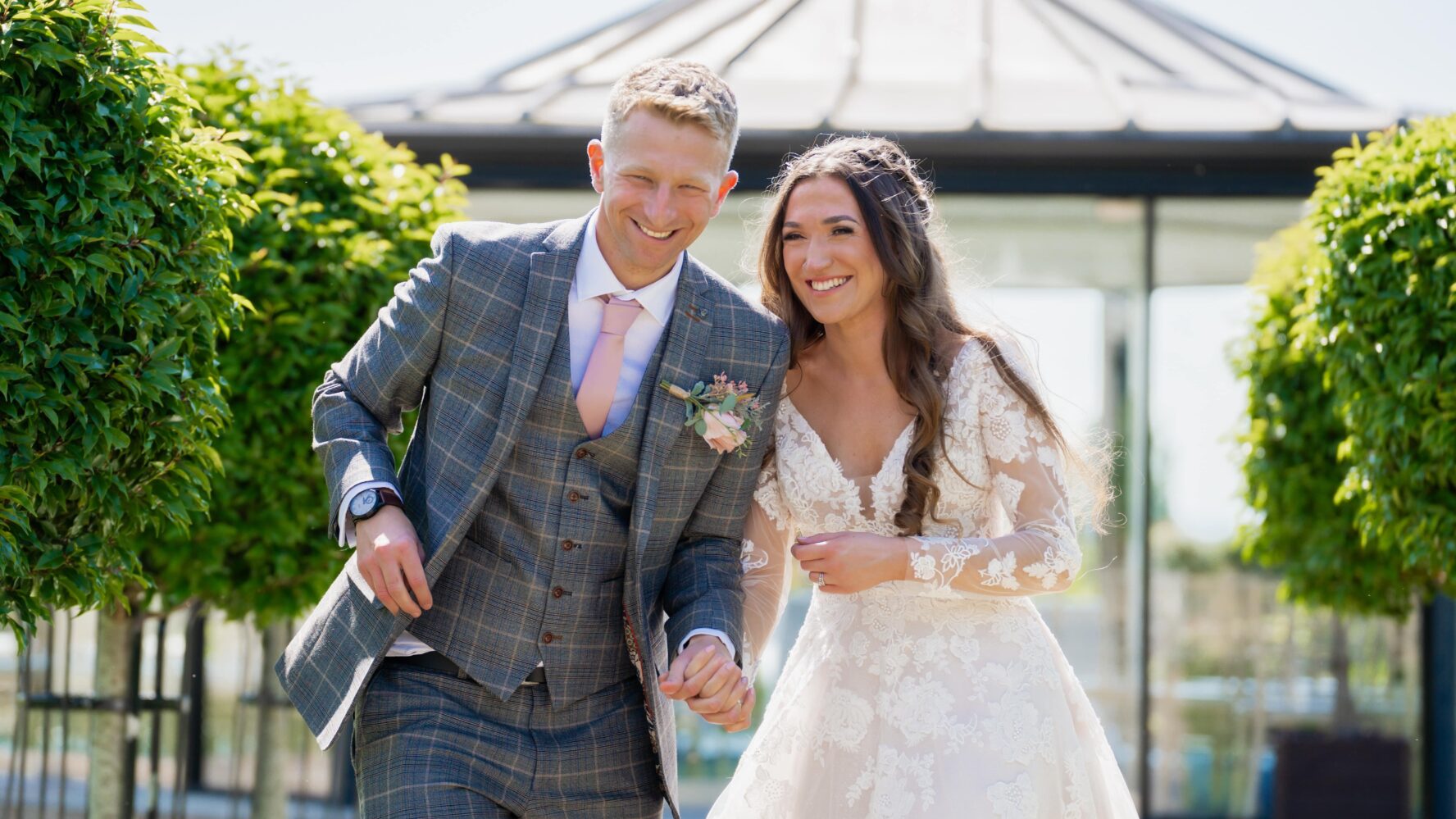 Joe,a white man with blonde short hair, with his wife Kathryn, a white lady with long, straight brown hair, on their wedding day.  Joe is wearing a grey checked suit with pale pink tie and Kathryn is wearing a long sleeveed white wedding dress. They both look delighted and are holding hands.