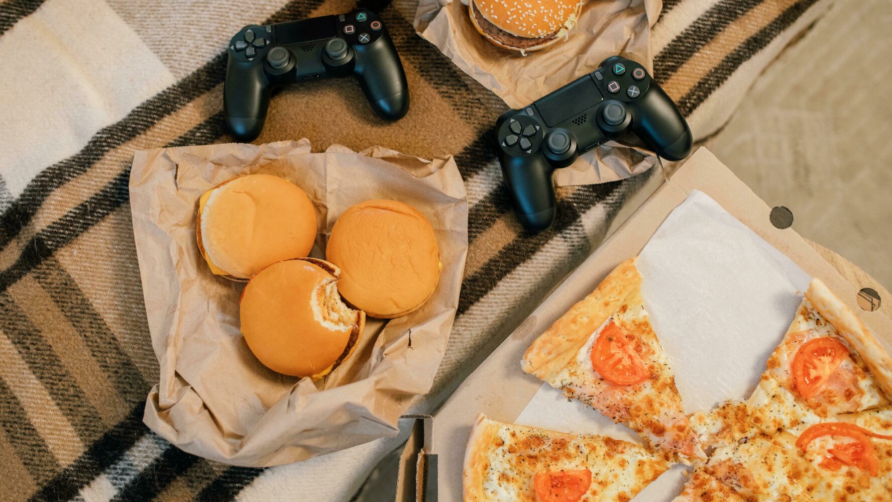 A takeaway and games night in: A beige tartan picnic blanket laid out with an assortment of pizza and bugers, with two games console controllers. Photography by Alena Darmel.