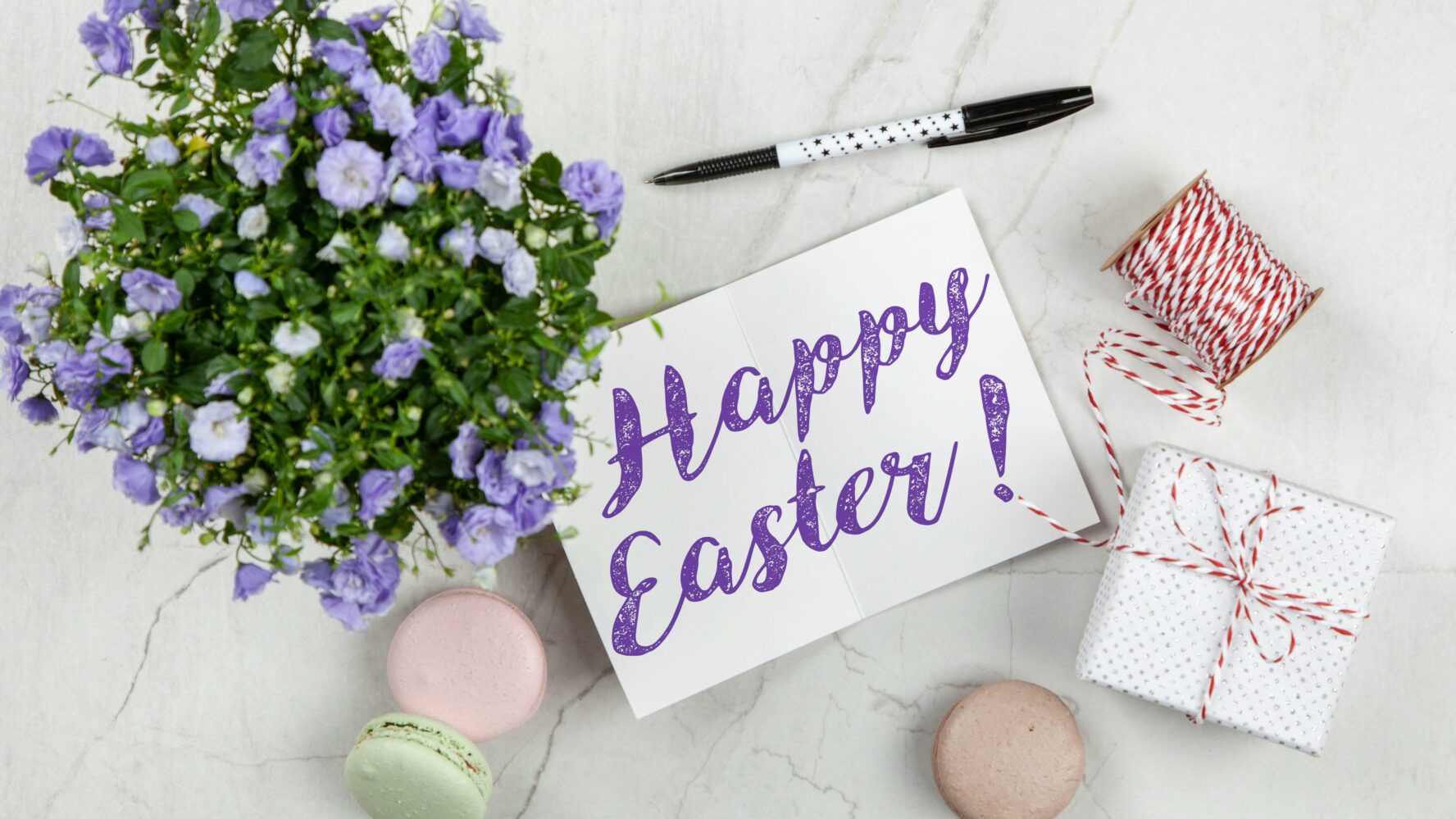 Image of a card that says 'Happy Easter' on the front, next to some purple flowers on a table, a pen, and a present