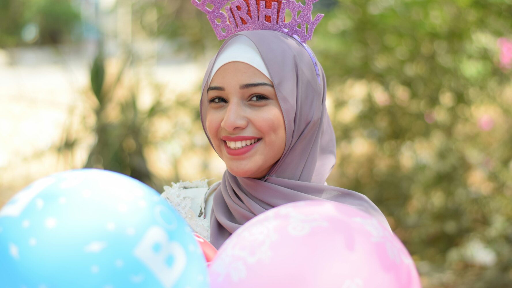 A young woman stood outside, smiling at the camera holding pink and blue balloons, wearing a birthday headband
