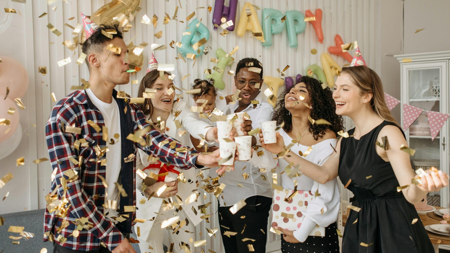 Friends gathered together at a party celebrating and smiling, stood together with confetti falling on them.