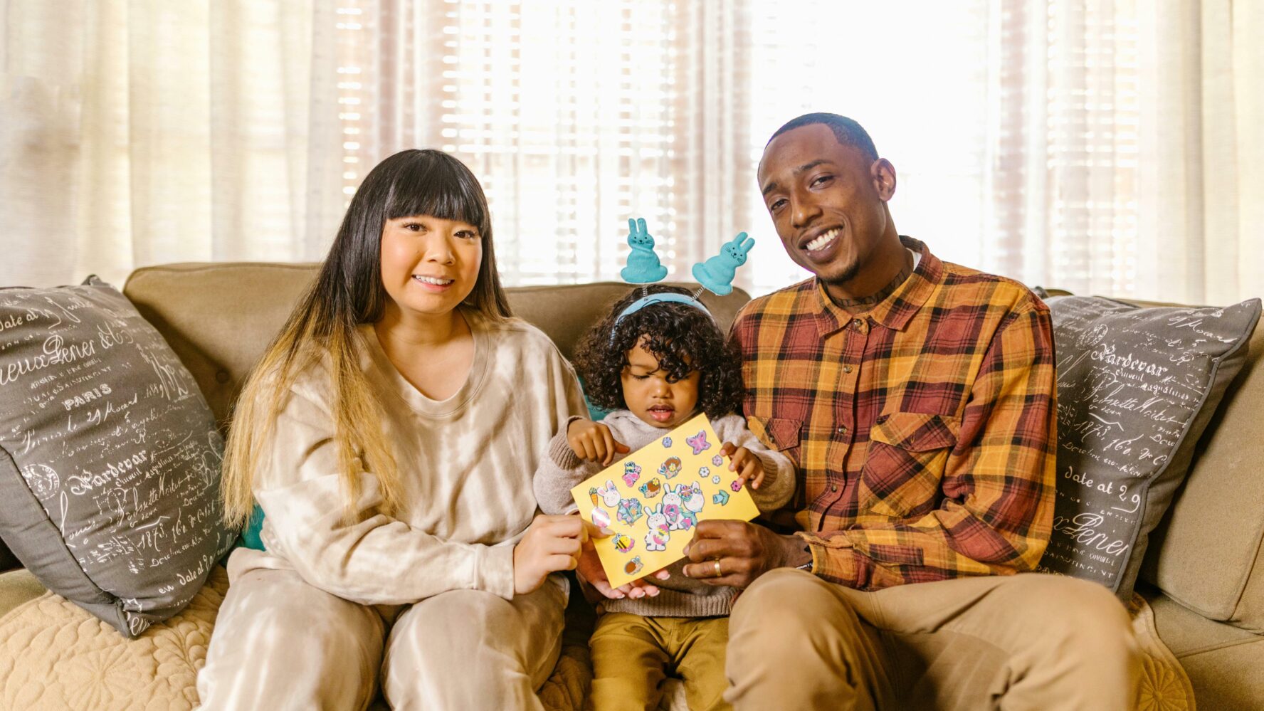 Image of two parents sat on their sofa with their toddler, smiling at the Easter card their daughter has made