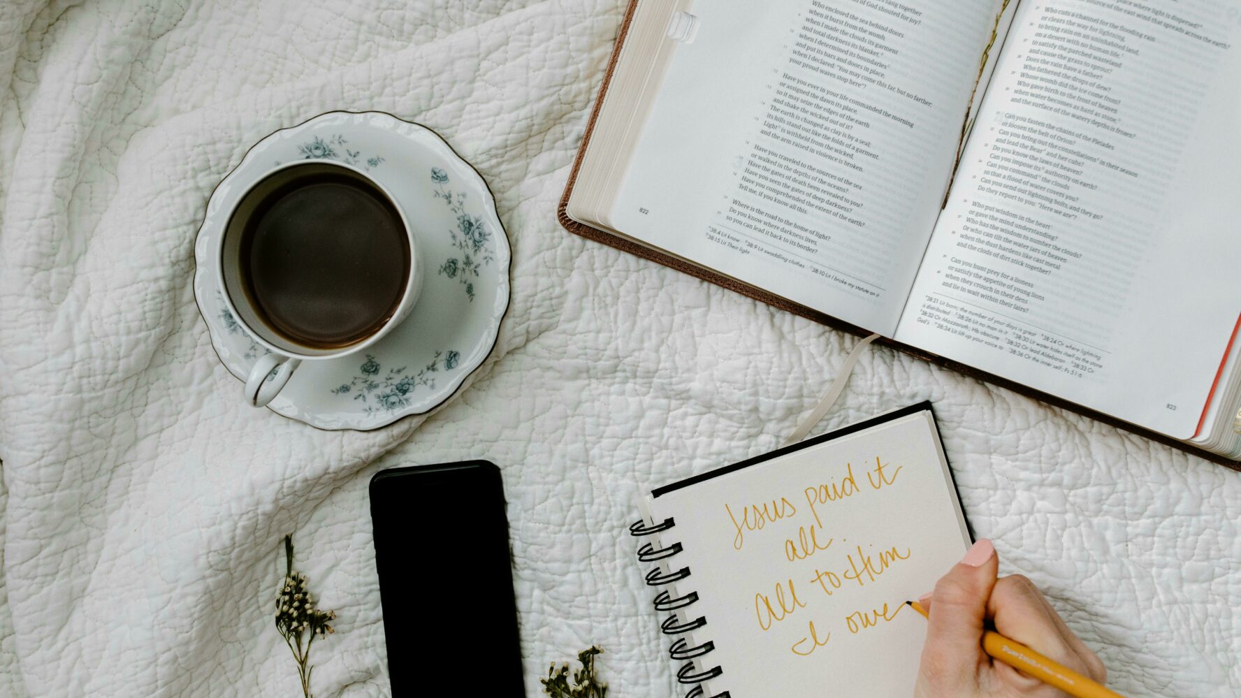 an open Bible, notebook, phone and tea cup set out on a white blanket