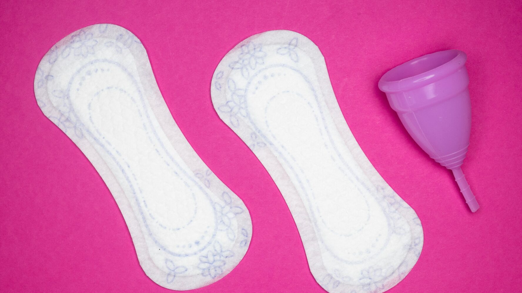 A pink background with period products on top, including two pads and a period cup