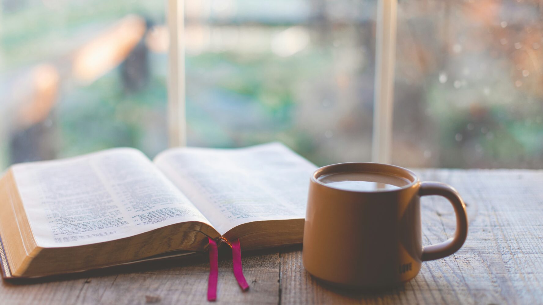A mug next to an open Bible on a wooden table, in front of a window overlooking a garden