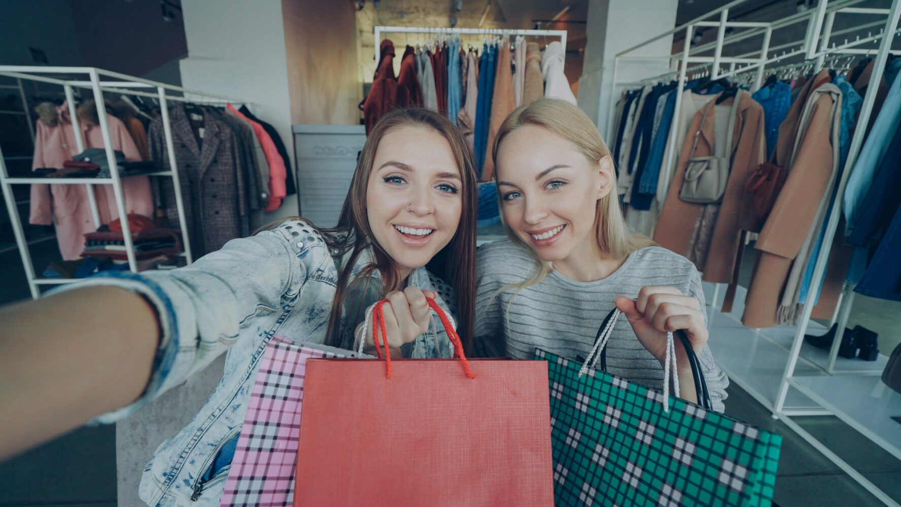 Two friends taking a selfie smiling, holding up shopping bags.