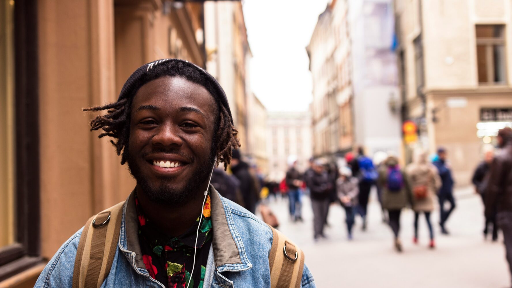 A young person wearing headphones and walking in a town centre, smiling at the camera