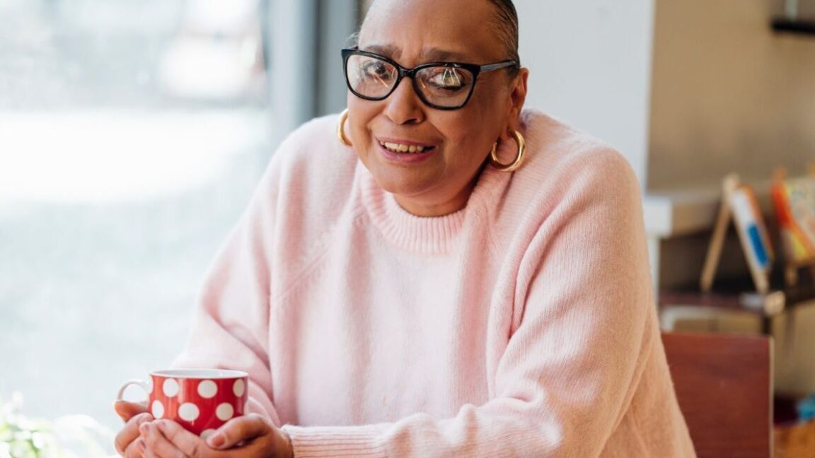 CAP client, Sharon, sat at a table holding a red mug, wearing a pink jumper and smiling at the camera